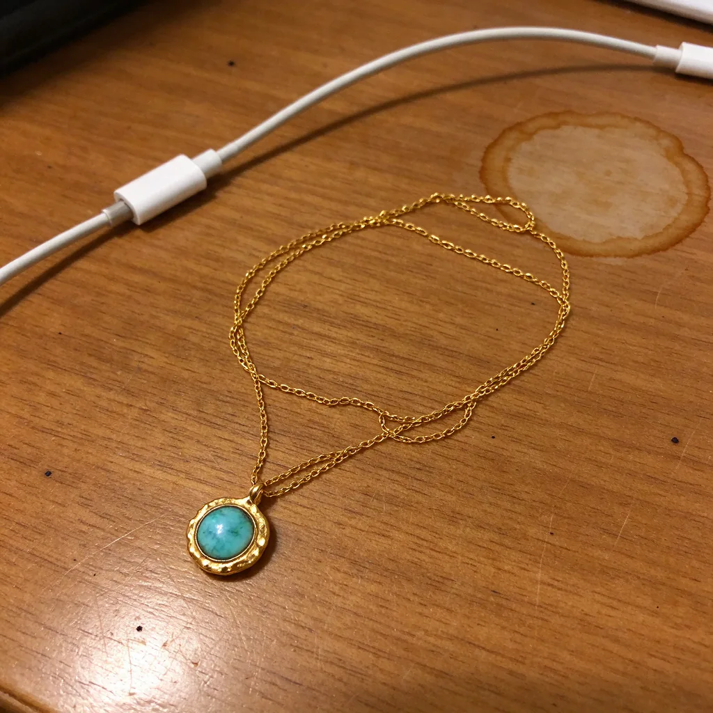 A gold pendant necklace with a turquoise stone lying tangled on a wooden desk.